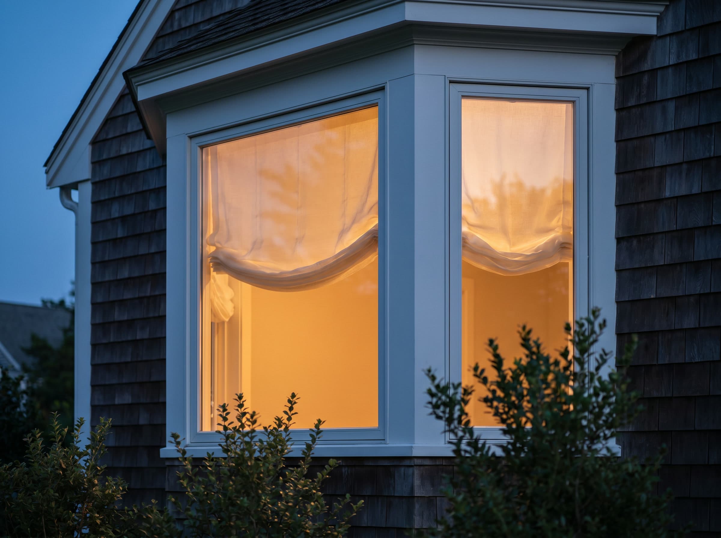 A New England home's bay window glowing at dusk through sheer shades.